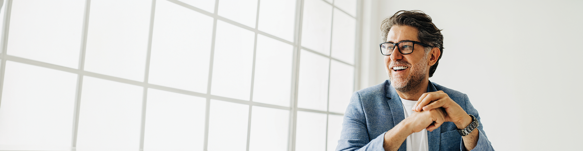 A smiling man with glasses sits in a bright, minimalist room, leaning on his hands near a large window with grid patterns.